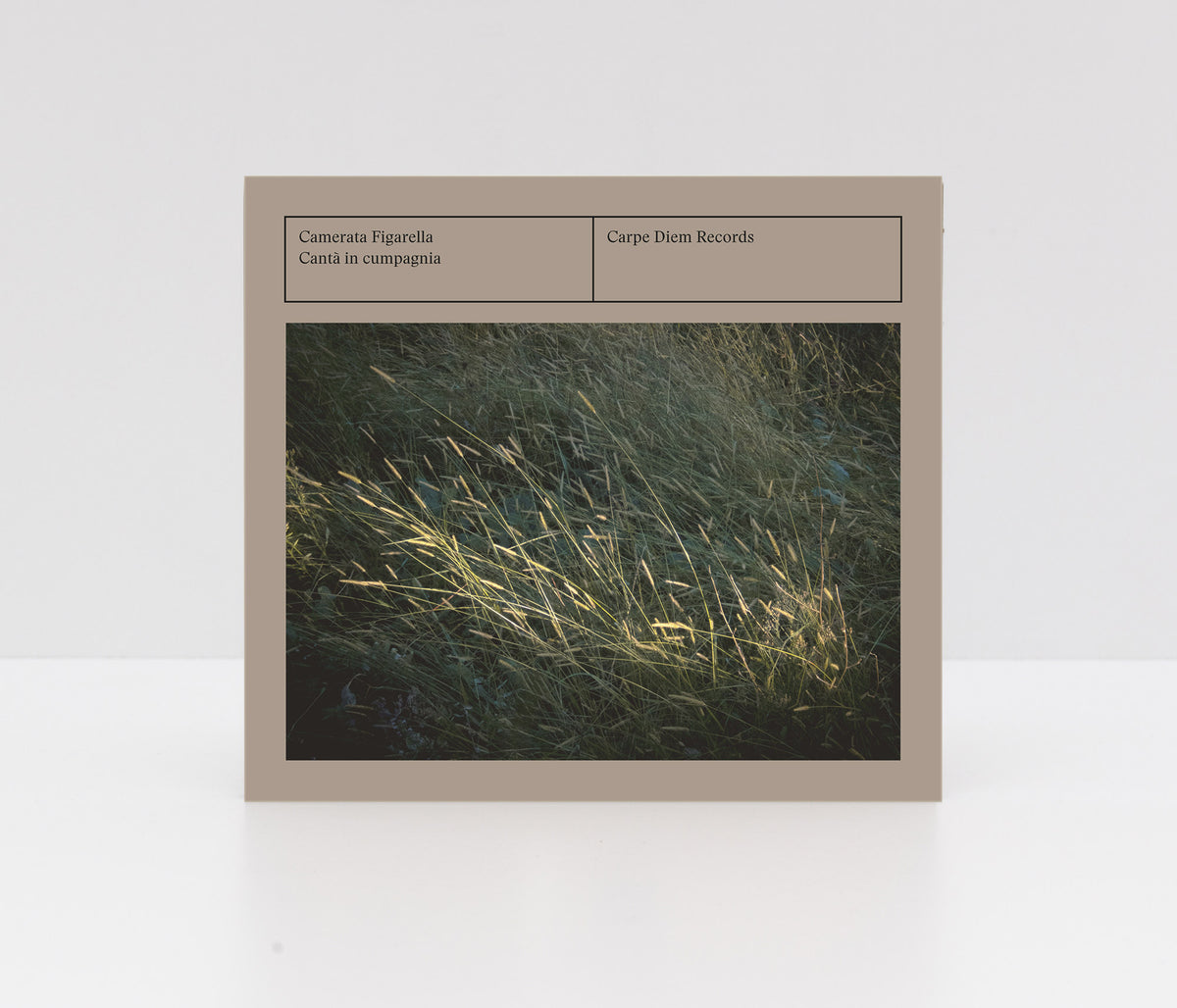 A square music album or book-style cover standing upright on a white surface against a white background. The cover has a warm taupe border with text reading 'Camerata Figarella : Cantà in cumpagnia' and 'Carpe Diem Records,' plus a central photograph of sunlit grasses in a dark field.