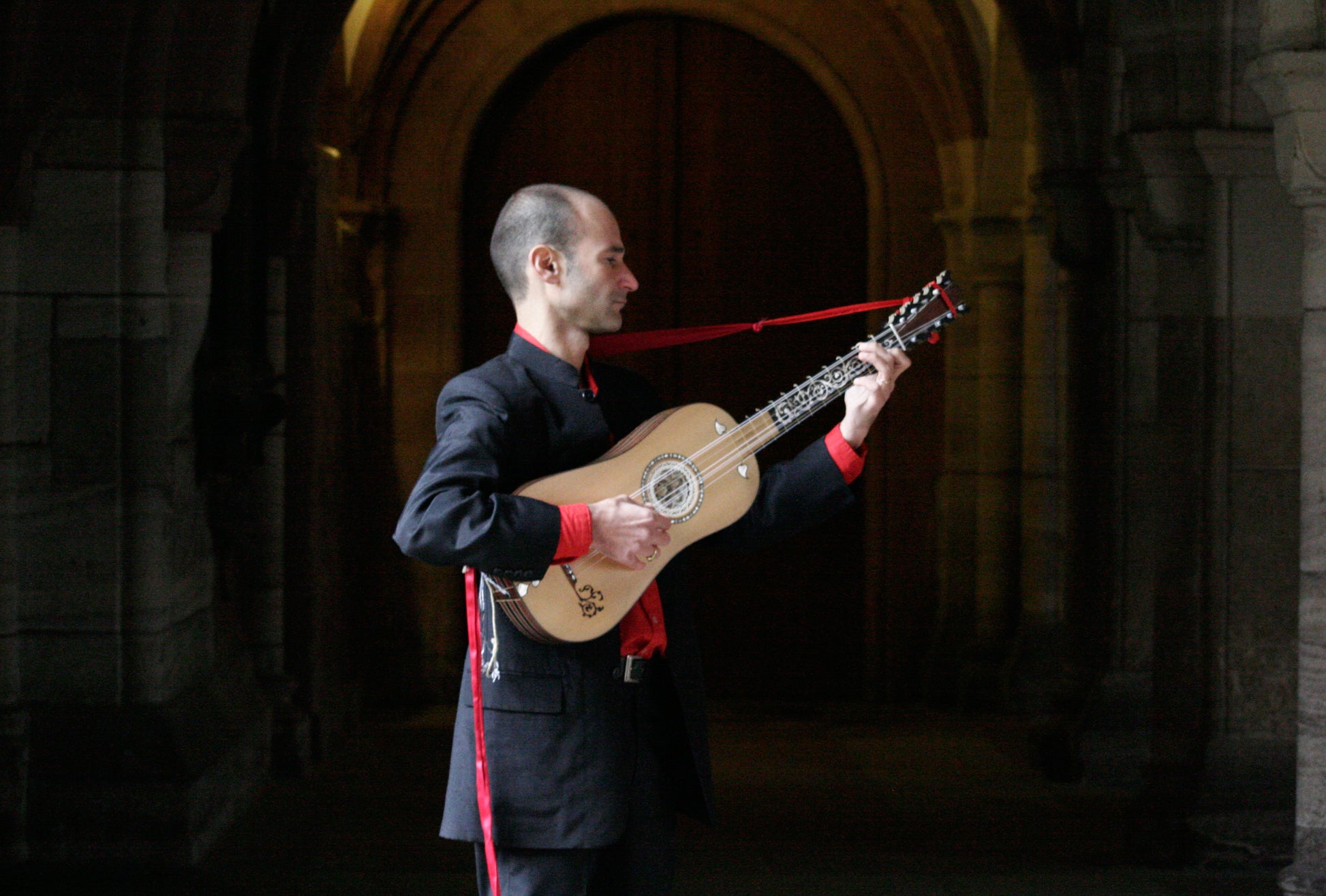 Rosario Conte playing a baroque guitar in front of a dark hallway in a castle. 